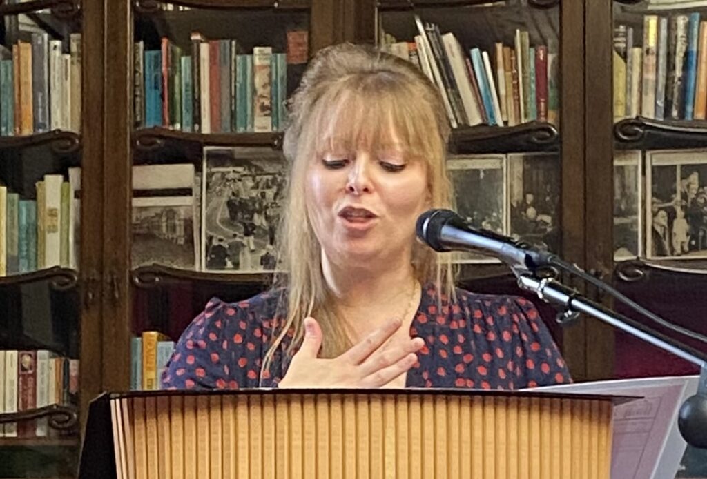 A woman with blonde hair in a messy bun stands behind a podium speaking into a microphone. She is wearing a black dress with red patterns, and is positioned in front of a cabinet full of books.